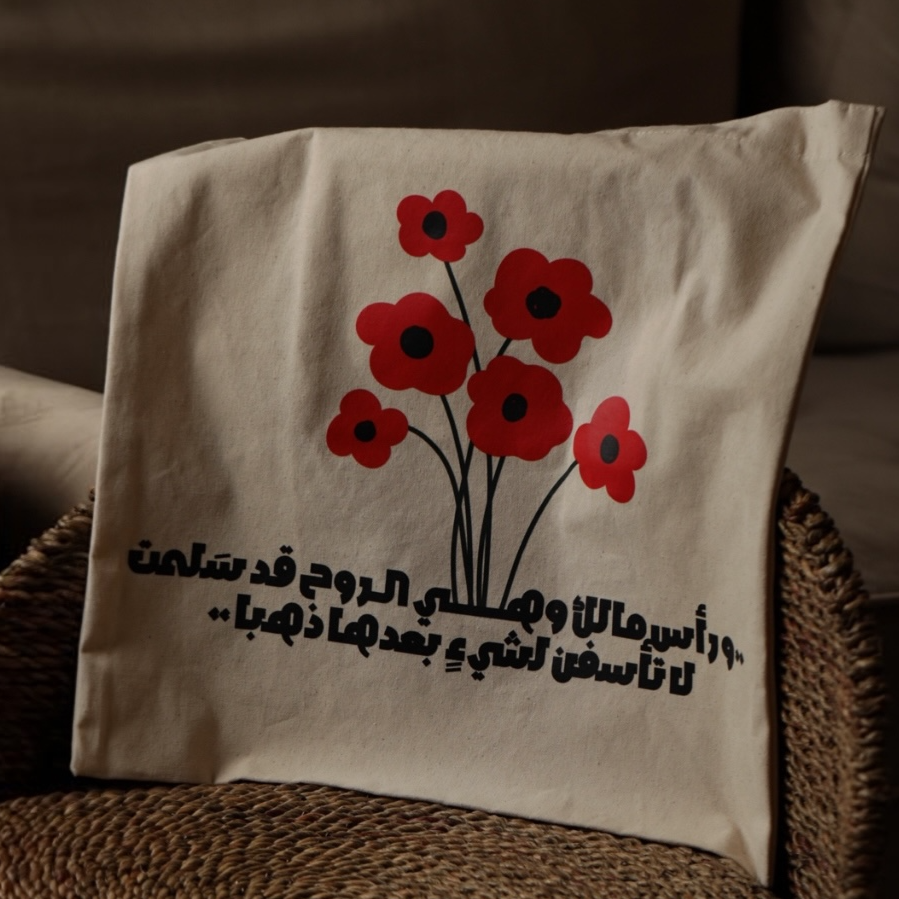 A cream-colored canvas throw pillow with a red poppy design and Arabic text, resting on a round woven brown chair in front of a beige sofa. A person is partially visible sitting on the sofa in the background.