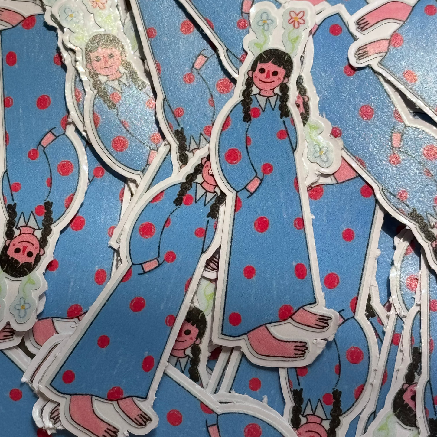 A pile of die-cut stickers featuring a cartoon girl with braided hair wearing a blue dress with red polka dots, shown on a light surface.
