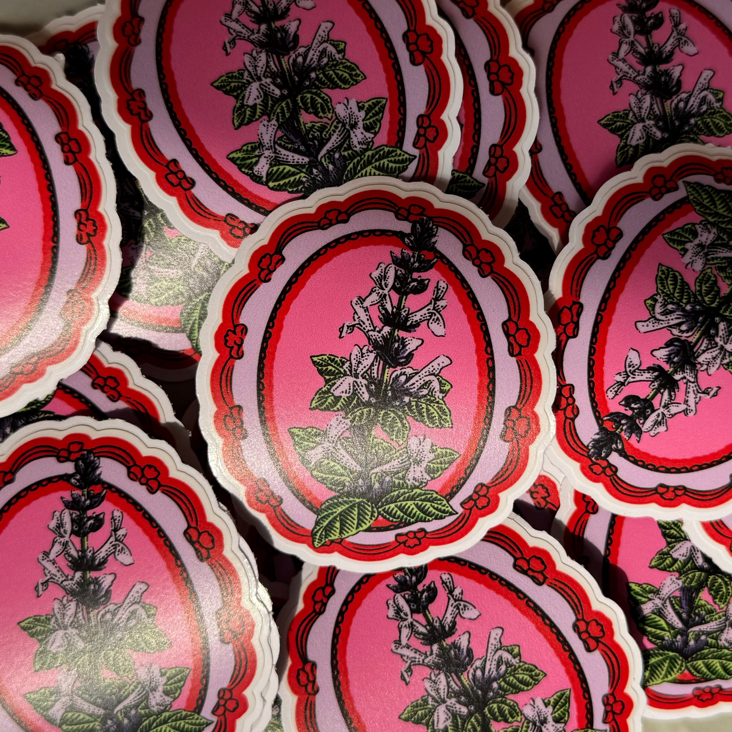 A close-up of multiple scalloped oval stickers or labels featuring a botanical illustration of a flowering plant in black, green, and white on a pink background with red ornamental borders.