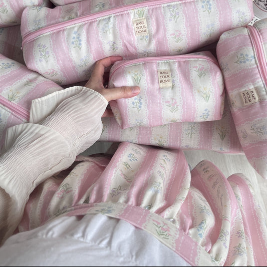 Pink and white striped fabric pencil cases with a small floral print and a visible 'The Pink Pig' label, arranged on a white surface with a hand holding one case. A mirror, flowers, and decorative props are visible in the background.