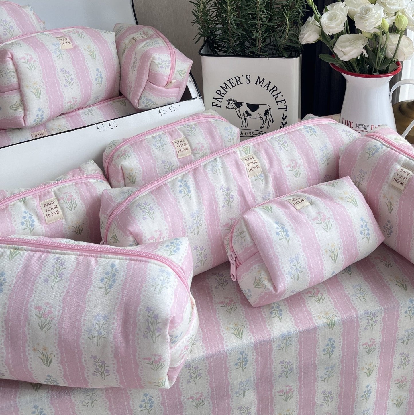 Pink and white striped bedding set with matching zippered pouches, styled on a bed in a bedroom setting with a lamp, flowers, and a plant.