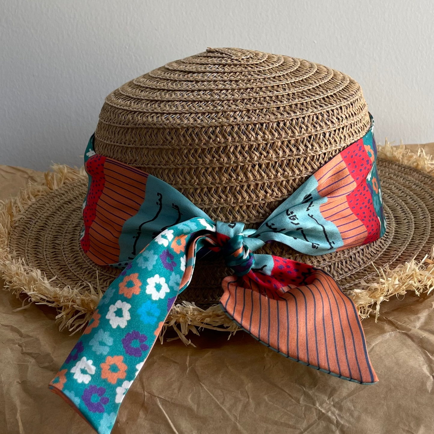 A woven straw sun hat with a wide brim, decorated with a colorful patterned silk ribbon tied in a bow around the crown. The ribbon features teal, red, orange, and white patterns.