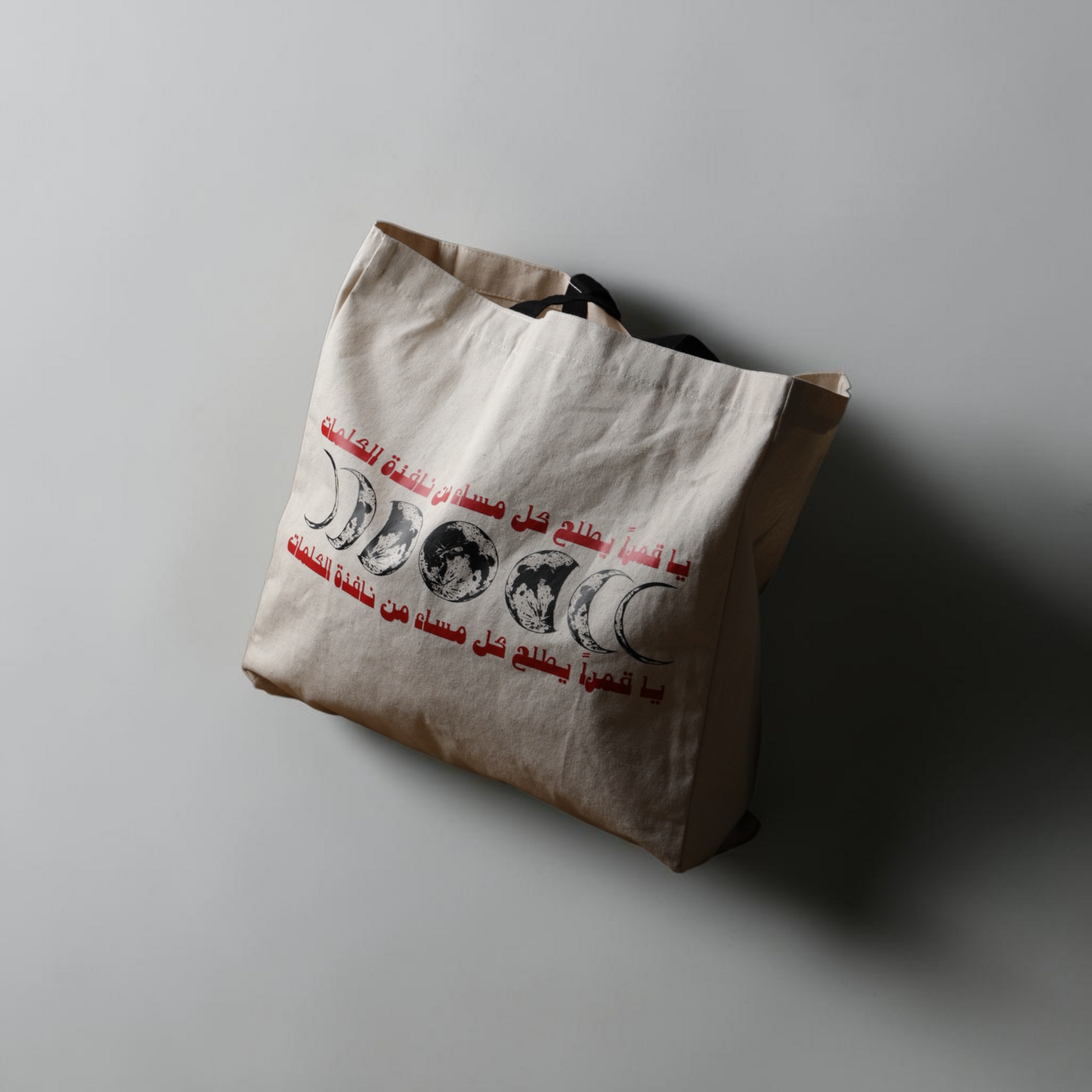 A beige canvas tote bag with black handles, featuring a red Arabic text print and a black-and-white moon phase graphic, resting on a gray surface with a soft shadow.
