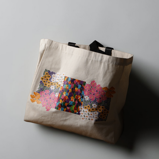 A beige canvas tote bag with black handles, featuring a colorful geometric and floral pattern printed on the front, set against a plain gray background with a soft shadow.