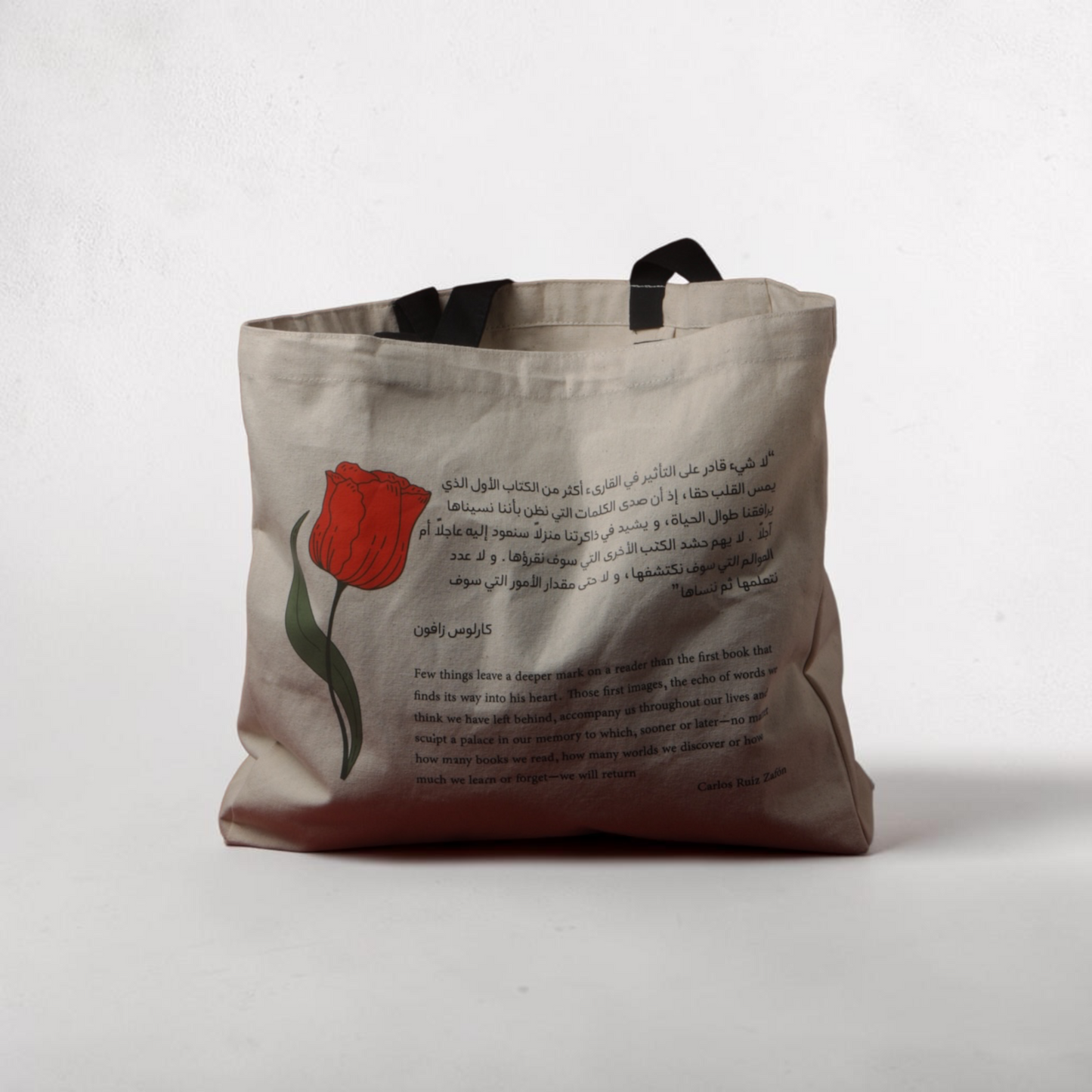 A canvas tote bag in a warm beige tone, featuring a red tulip illustration with green leaves and black Arabic text printed on the front. The bag has black fabric handles and is photographed against a neutral, light gray textured background.